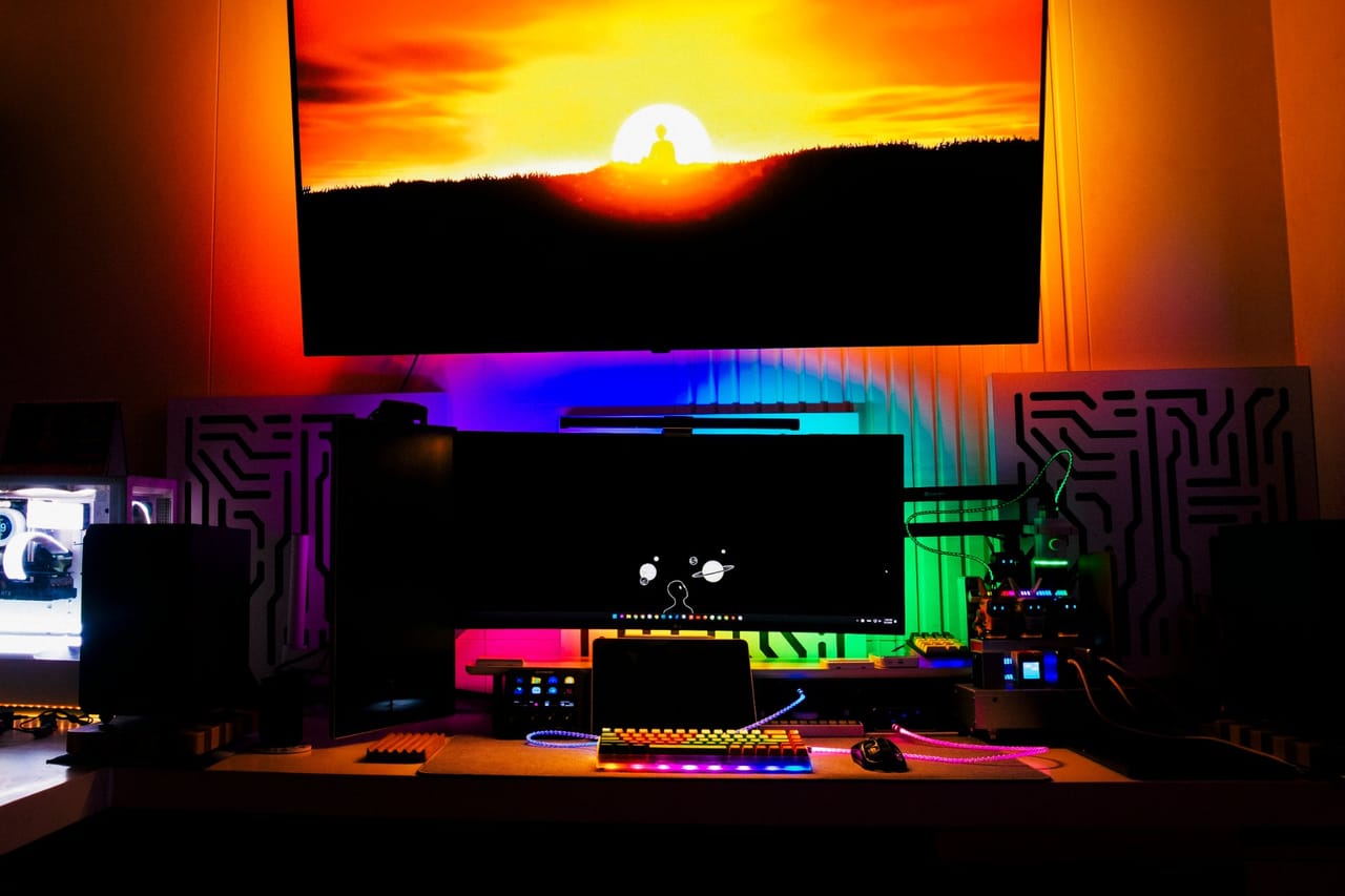 Góc máy của VGSer 9950: Góc máy tối với đèn RGB nhiều màu rực rỡ, TV lớn treo tường phía trên, màn hình ultrawide và bàn phím phát sáng bên dưới.
