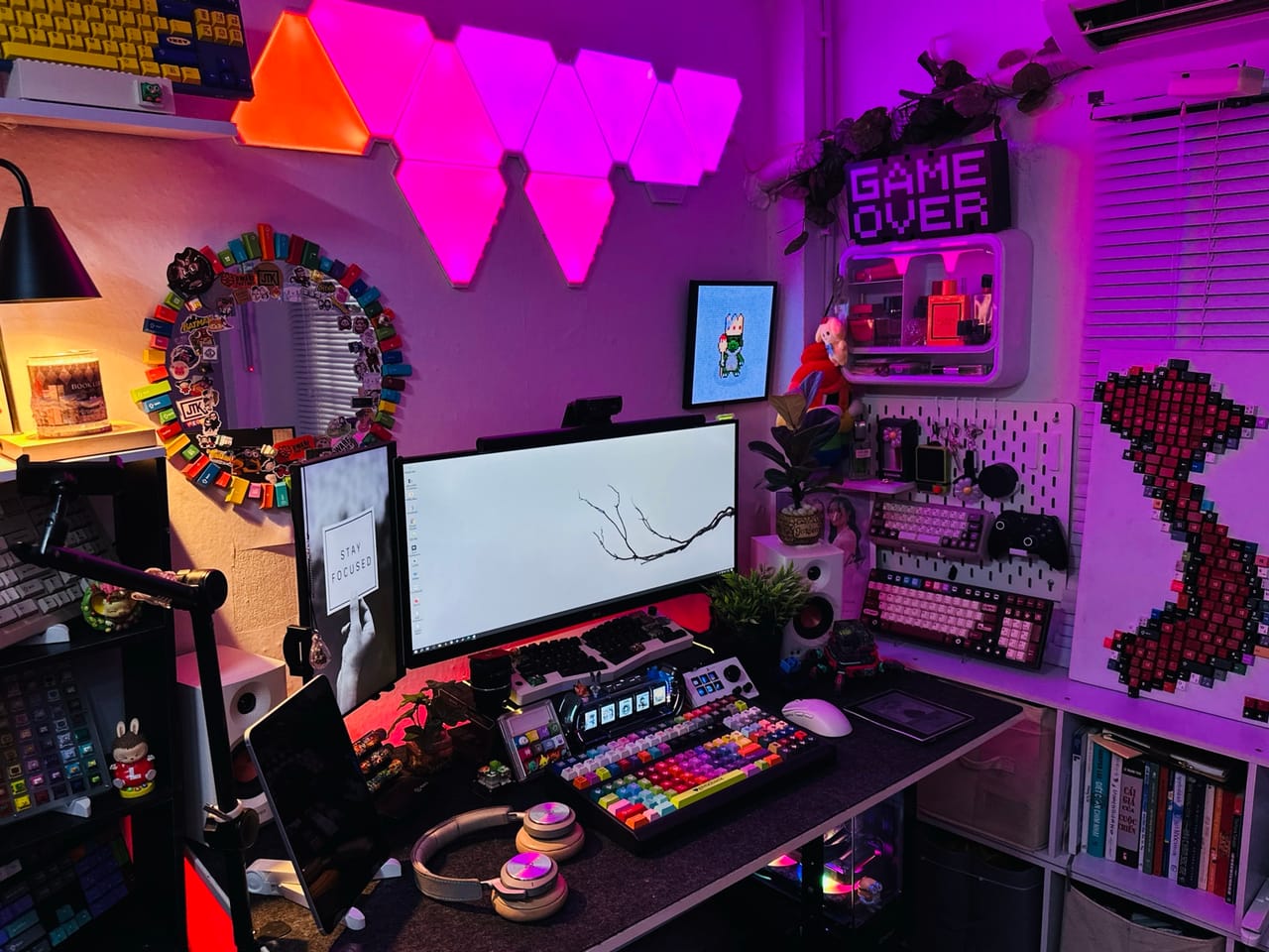 Góc máy của Thư Đoàn: Góc setup ánh đèn hồng tím neon với màn hình ultrawide, nhiều bàn phím custom keycap nhiều màu, bảng pegboard và tranh ghép keycap trên tường.