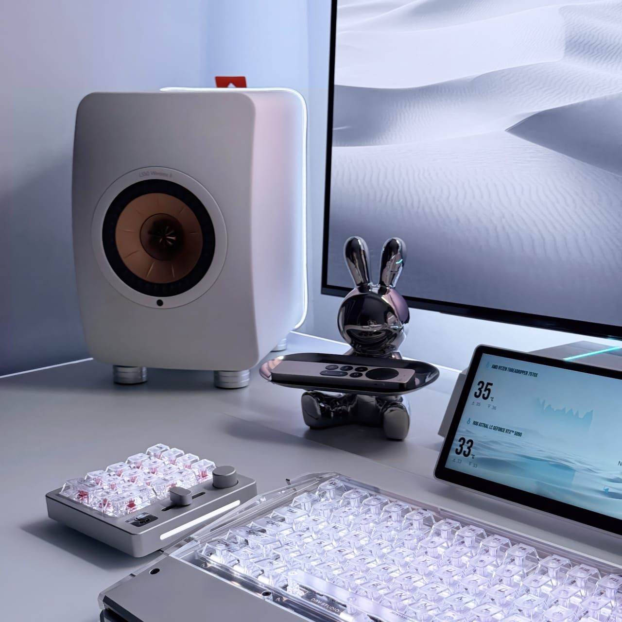 Góc máy của TaeLee: Góc bàn tông trắng bạc tối giản với loa KEF, bàn phím trong suốt, macro pad, màn hình phụ hiển thị nhiệt độ và tượng thỏ crom.