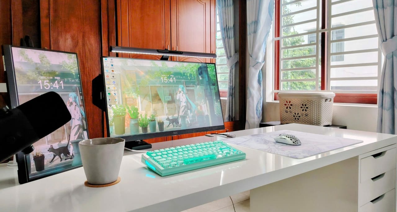 Góc máy của Zéphyr: Góc máy sáng sủa bên cửa sổ với bàn trắng, bàn phím xanh mint, chuột trắng, hai màn hình hình nền anime và đèn treo màn hình.