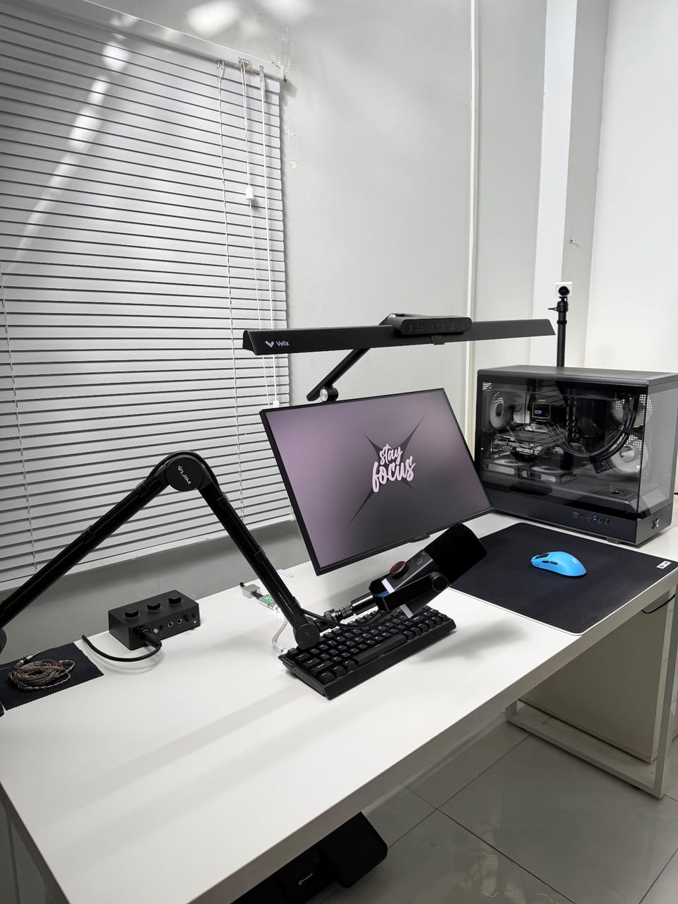 Setup góc máy của Intdire: Góc bàn trắng gọn gàng với màn hình, đèn treo màn hình, micro gắn arm, bàn phím đen, chuột xanh và PC case kính.