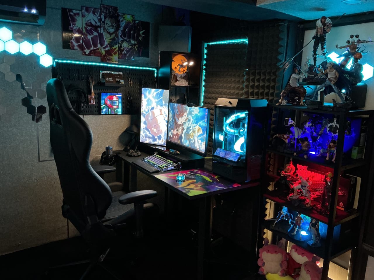 Góc máy của Rookie Bùi: Góc máy tối với đèn LED xanh, hai màn hình hình anime, PC case kính, kệ trưng bày figure phát sáng đỏ xanh và tường tiêu âm.