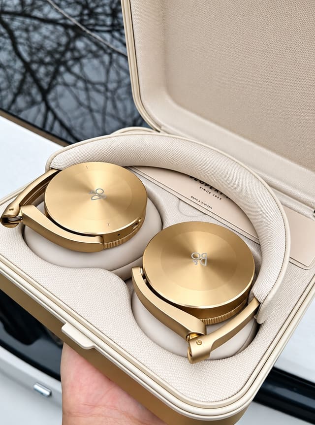 Cặp tai nghe over-ear màu vàng gold của Bang & Olufsen nằm trong hộp đựng màu be sang trọng, chụp ngoài trời.
