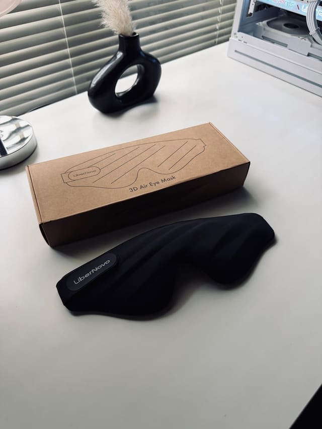 Hộp và chiếc bịt mắt 3D Air Eye Mask của LiberNovo đặt trên bàn trắng, cạnh bình hoa đen và một case PC trắng phía sau.