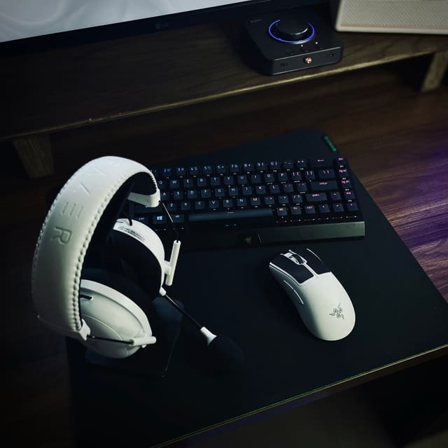 Góc bàn gỗ tối màu với tai nghe và chuột Razer màu trắng nổi bật cạnh bàn phím đen trên tấm lót chuột lớn.