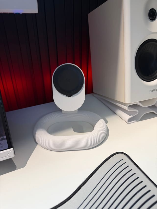 Góc bàn trắng tối giản với loa Edifier, webcam gắn đế vân sóng, lót chuột sọc và nền rèm đỏ đen phía sau.