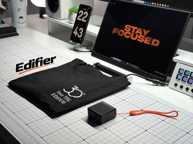 Góc bàn trắng gọn gàng với loa bluetooth mini Edifier, áo thun kỷ niệm 30 năm, màn hình hiển thị Stay Focused và bàn phím điều khiển.