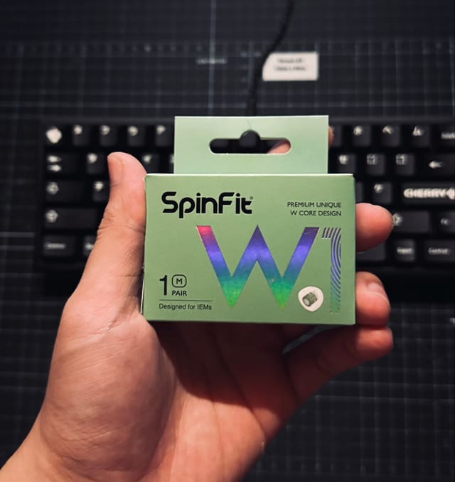 Hộp đệm tai nghe SpinFit W1 màu xanh mint được cầm trước bàn phím đen Cherry đặt trên tấm lót bàn tối màu.