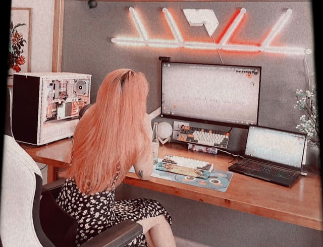 Góc setup tông trắng hồng pastel với bàn gỗ, PC tản nhiệt nước, hai bàn phím cơ, laptop và đèn panel LED cam trên tường.
