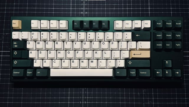 Bàn phím cơ TKL vỏ xanh lá đậm với keycap trắng và phím be nhấn màu, đặt trên tấm lót cắt dạng lưới tối màu.