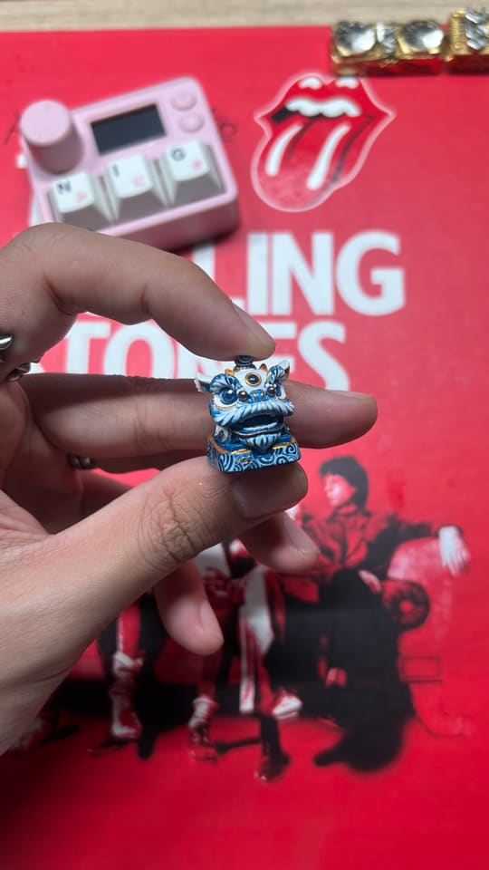 Cận cảnh một chiếc keycap artisan hình lân xanh trắng được cầm trên tay, phía sau là macropad hồng và tấm lót đỏ Rolling Stones.