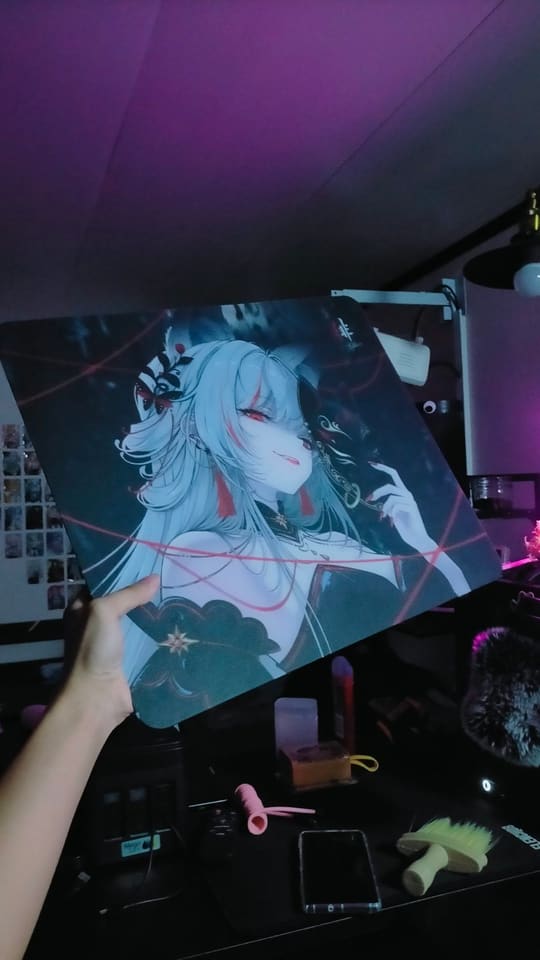 Một tấm lót chuột cỡ lớn in hình nhân vật anime tóc bạc được giơ lên trước bàn có micro, điện thoại và ánh đèn tím.