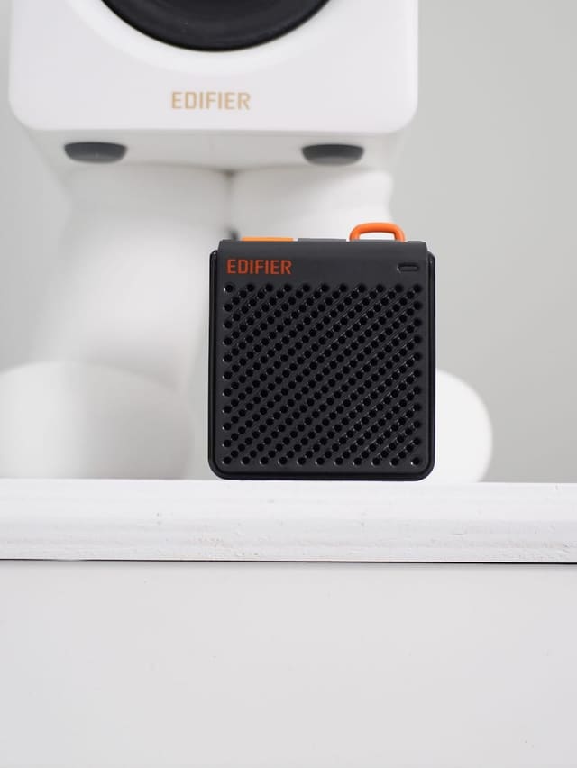 Loa bluetooth mini màu đen điểm cam của Edifier đặt trên kệ trắng, phía sau là chiếc loa Edifier trắng hình robot.