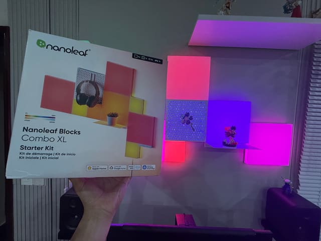 Hộp Nanoleaf Blocks Combo XL được giơ lên trước bộ đèn panel treo tường phát sáng hồng tím, bên cạnh có loa và mô hình nhân vật.