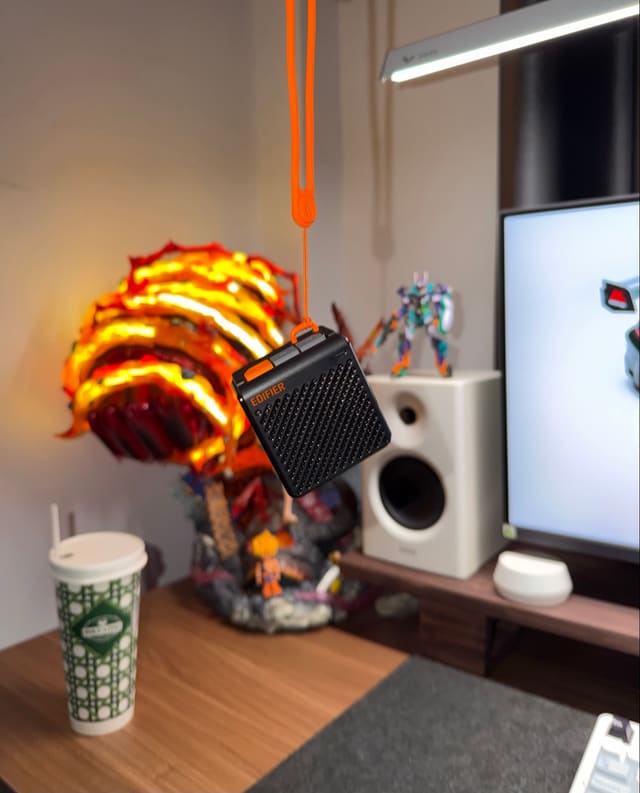 Loa bluetooth Edifier nhỏ màu đen cam treo bằng dây đeo, phía sau là mô hình anime phát sáng, loa trắng và màn hình trên bàn gỗ.