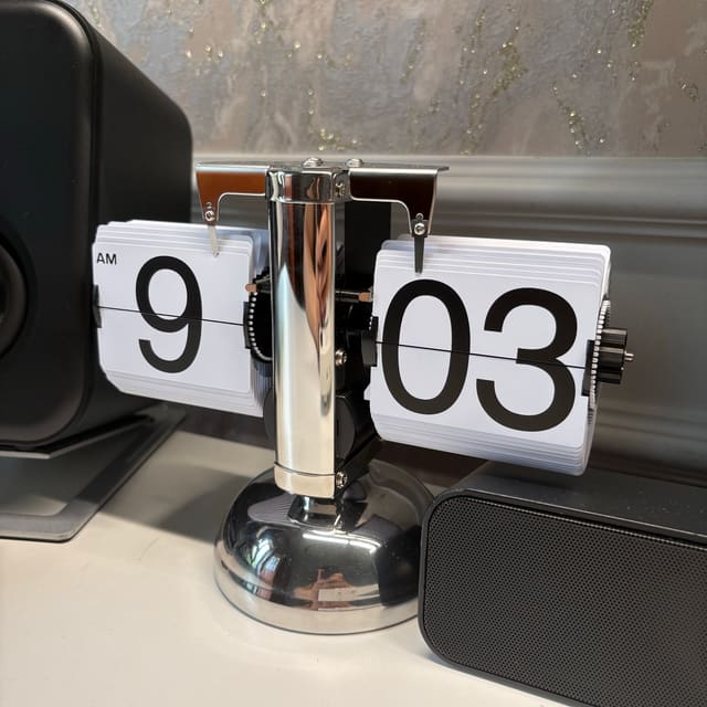 Đồng hồ lật phong cách retro bằng inox bóng đặt giữa hai chiếc loa đen trên mặt bàn trắng, hiển thị 9:03 AM.