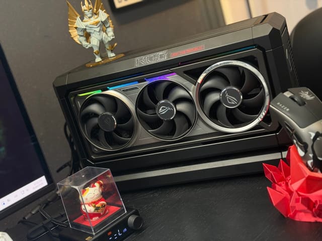 Góc bàn màu đen nổi bật chiếc card đồ họa ROG đèn RGB, bên cạnh là chuột Razer, tượng mèo may mắn và mô hình trang trí.
