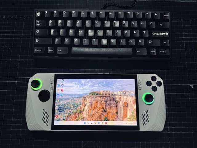 Bàn phím Cherry 60% keycap tiếng Nhật đặt cạnh máy chơi game cầm tay màu trắng có joystick sáng xanh lá trên nền thảm tối.