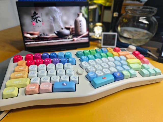 Bàn phím công thái học màu trắng kem với keycap cầu vồng nhiều màu đặt trên mặt bàn vàng, phía sau là màn hình hình nền trà đạo.
