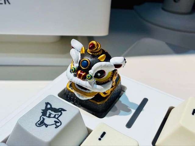 Cận cảnh keycap artisan hình lân múa đen trắng viền vàng trên bàn phím cơ trắng kem, bên cạnh là keycap xám hình chú chó.