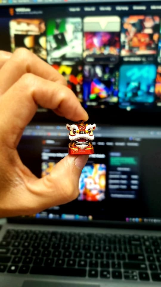 Cận cảnh keycap nghệ thuật hình đầu lân đỏ trắng được cầm trước laptop và màn hình đang mở trang web nền tối.